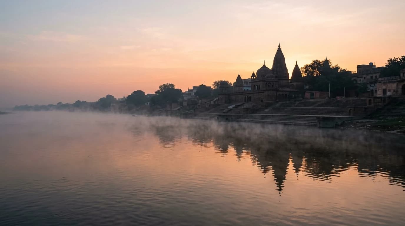 The Yamuna river at dawn with low mist hovering above the water, a distant ghat and silhouetted temple, soft amber-pink sky.